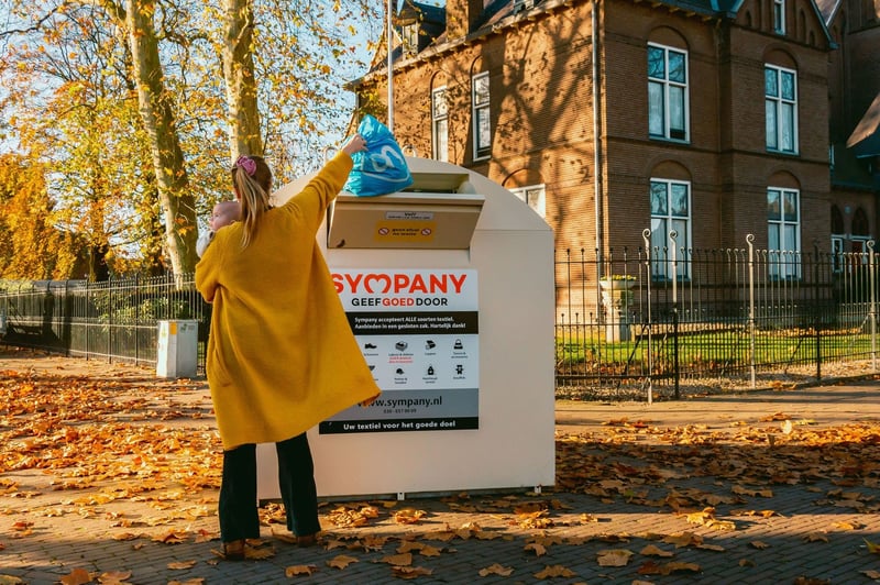 Vrouw in gele trui gooit een blauwe plastic tas in een grote witte inzamelingscontainer voor textiel.